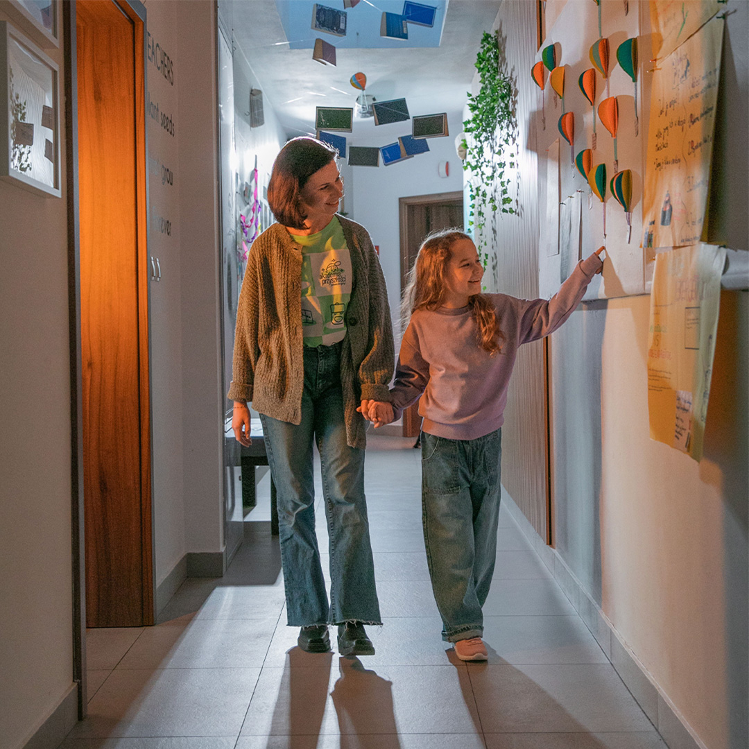 A girl holds hands with a woman as they walk through a corridor looking at artwork on the wall.